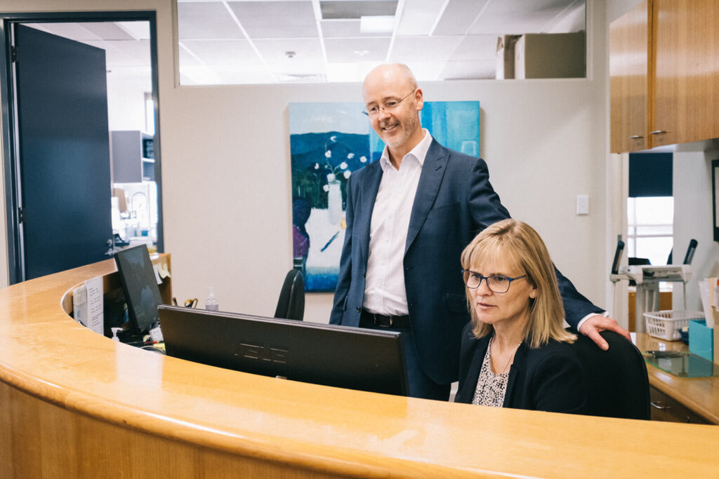 A man in a suit stands next to a seated woman at a reception desk, both looking at a computer monitor in an office environment.