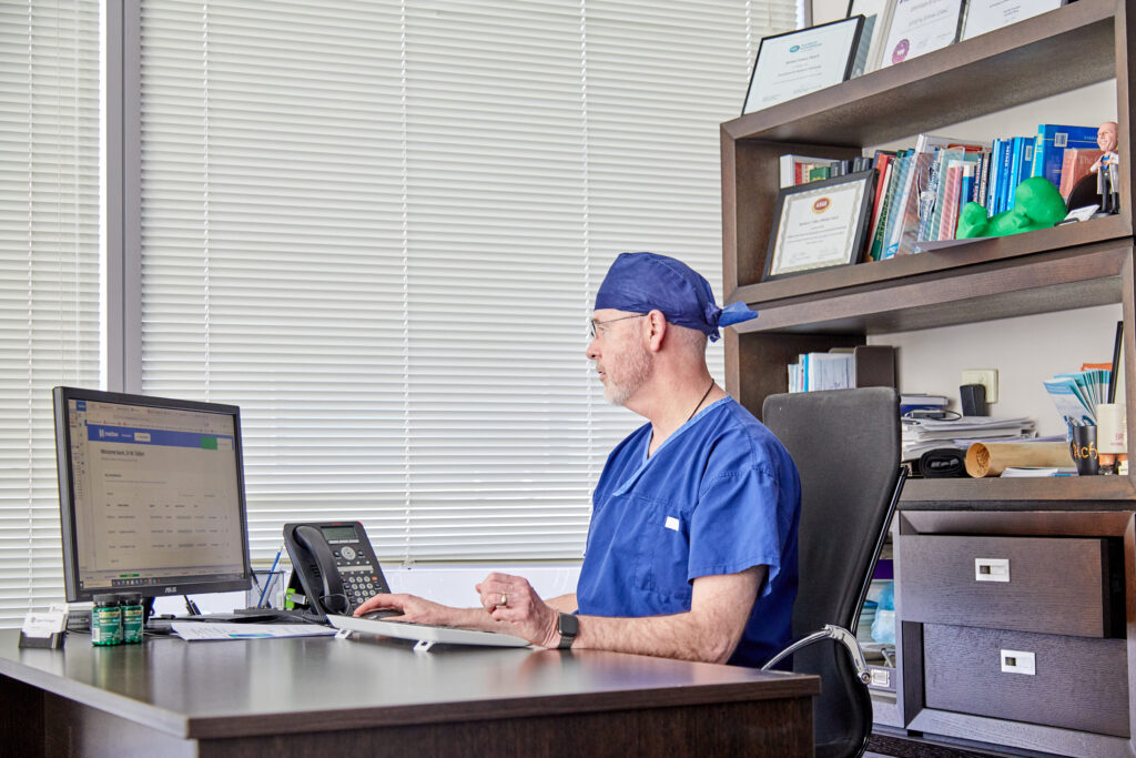 A medical professional in blue scrubs sits at a desk, using a computer in an office with shelves of books and certificates on the wall.