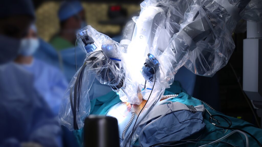 Robotic surgical system with mechanical arms performing a procedure on a patient in an operating room, with medical staff present in the background.